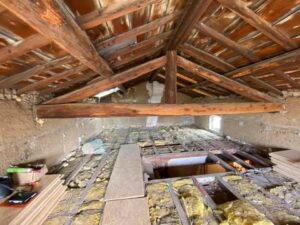 Grenier en rénovation : poutres bois apparentes, isolation jaune entre solives du plancher, planches neuves. Murs bruts, petite fenêtre.