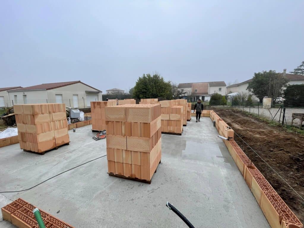 Vue de chantier : homme sur dalle béton. Piles de briques de terre cuite et début de murs périphériques, maisons en arrière-plan.