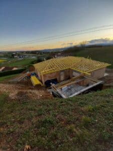 Maison en construction avec charpente bois au coucher du soleil. Piscine ou sous-sol en gros œuvre sur terrain vallonné.