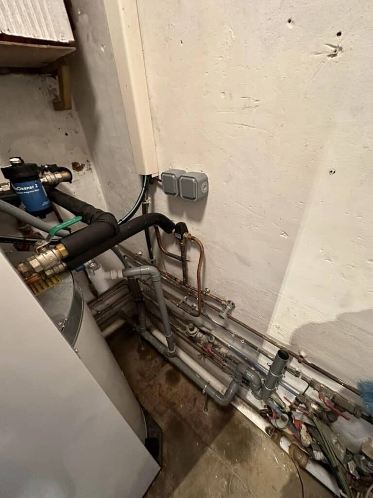 Installation technique de chaufferie avec filtre magnétique Cleaner 2, chauffe-eau, diverses tuyauteries isolées, prises électriques murales et sol sale.