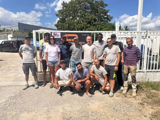 Équipe de travail posant devant le site DPLSS Groupe d'environ 12 personnes, hommes et femmes, posant devant une clôture blanche avec un logo orange flou, sous un ciel bleu et ensoleillé.