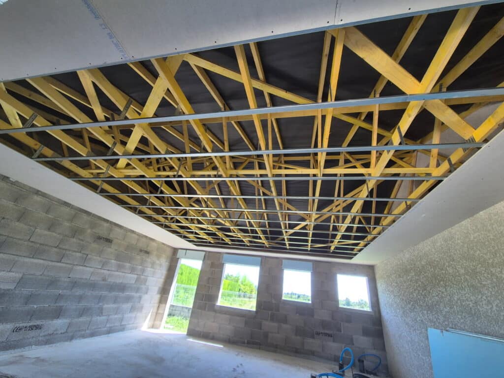 Construction d'un plafond suspendu avec ossature métallique sous charpente bois et pare-pluie noir, murs en parpaings et fenêtres.