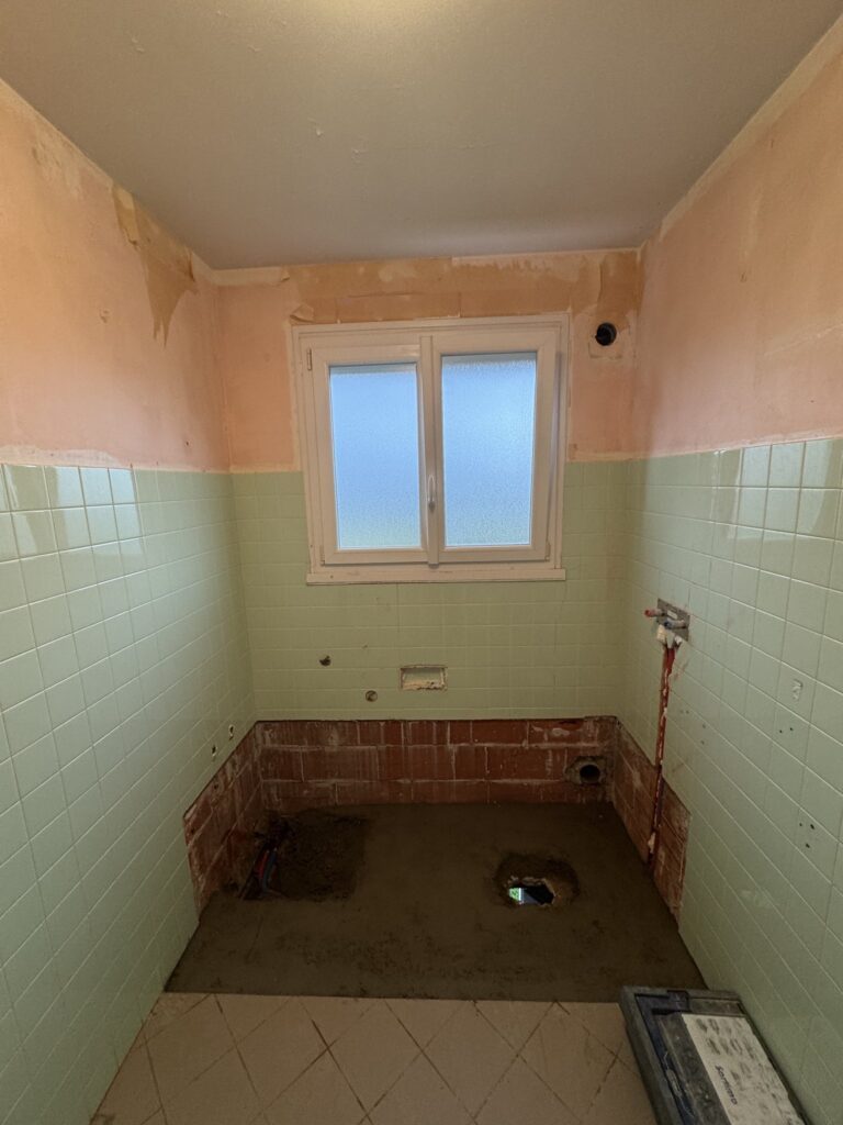 Petite salle de bain en cours de rénovation avec carrelage vert, fenêtre et sol en béton pour plomberie.
