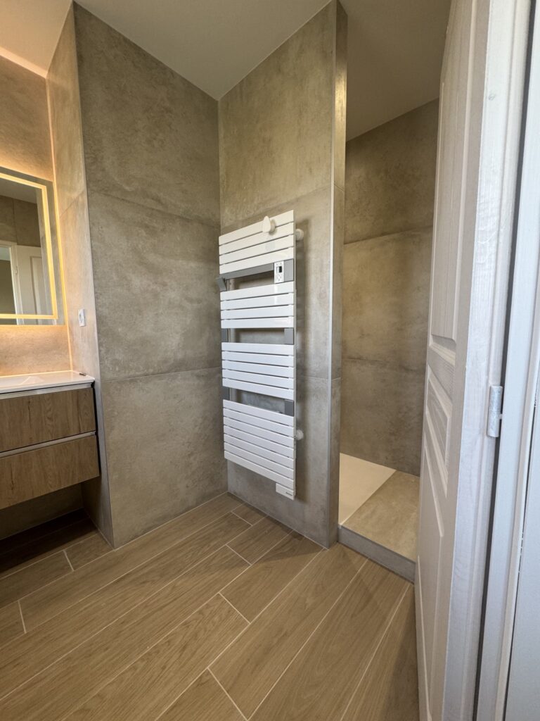 Salle de bain moderne avec carrelage effet pierre, sèche-serviettes blanc, meuble vasque en bois et douche à l'italienne.