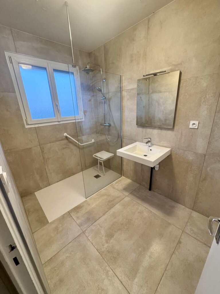 Salle de bain moderne avec douche à l'italienne accessible, siège de douche, lavabo suspendu et carrelage beige.