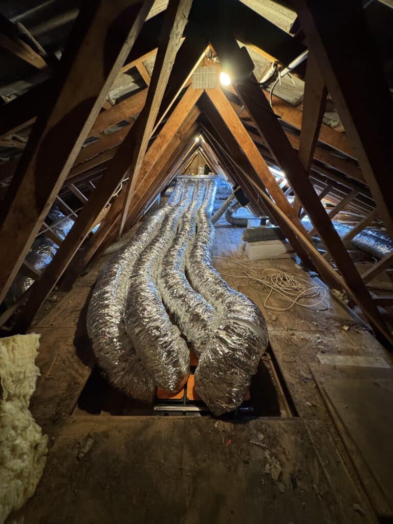 Plusieurs conduits d'air isolés argentés serpentant sous la charpente en bois d'un grenier sombre.